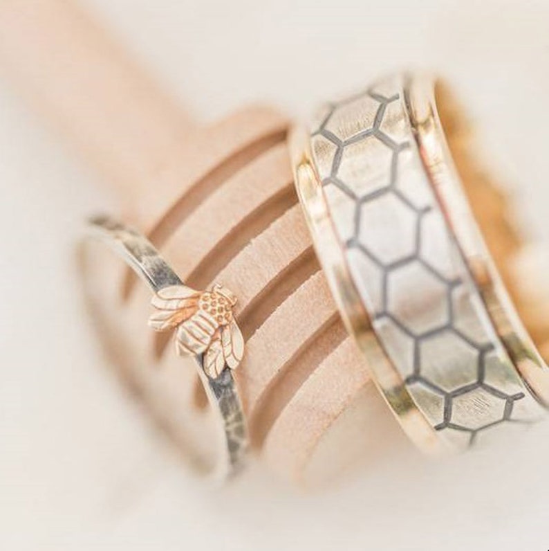 May include: Two wedding rings. One ring has a gold bee charm. The other ring has a gold band with a silver honeycomb pattern. The rings are resting on a wooden honey dipper.
