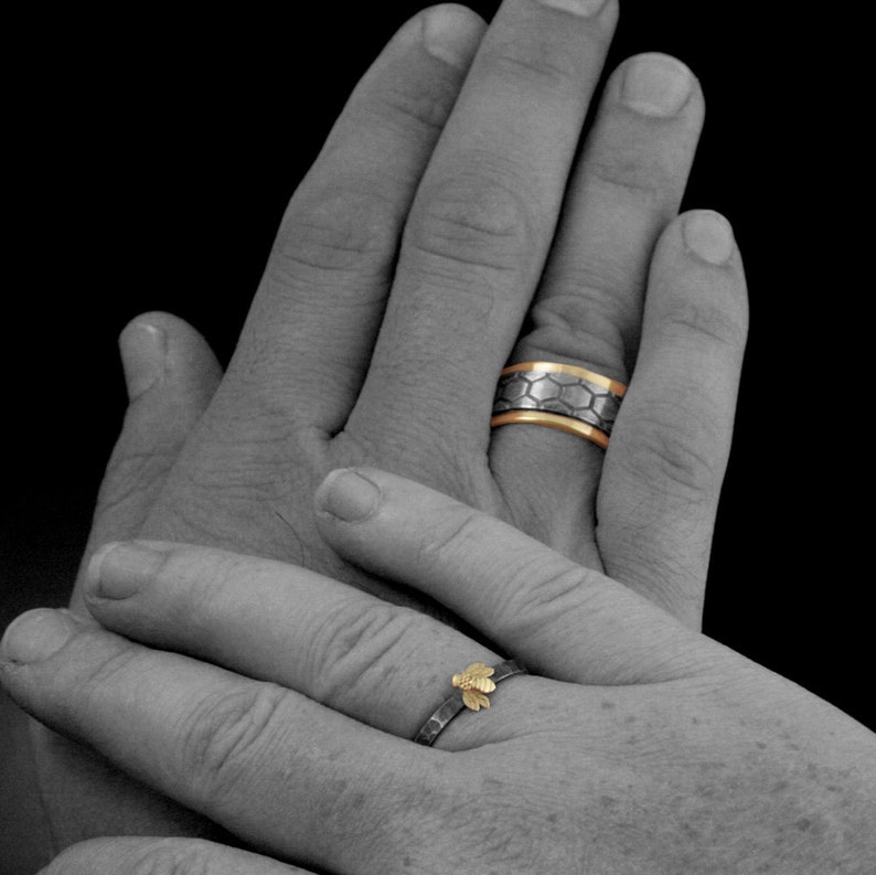 May include: Two wedding rings, one with a honeycomb design and gold accents, the other with a small gold bee. The rings are on a person's hand, with a black background. The rings are likely made of metal.