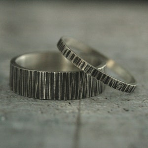 May include: Two silver rings with a textured, bark-like pattern. One ring is wider than the other, and both have a dark, oxidized finish. The rings are set against a gray background.