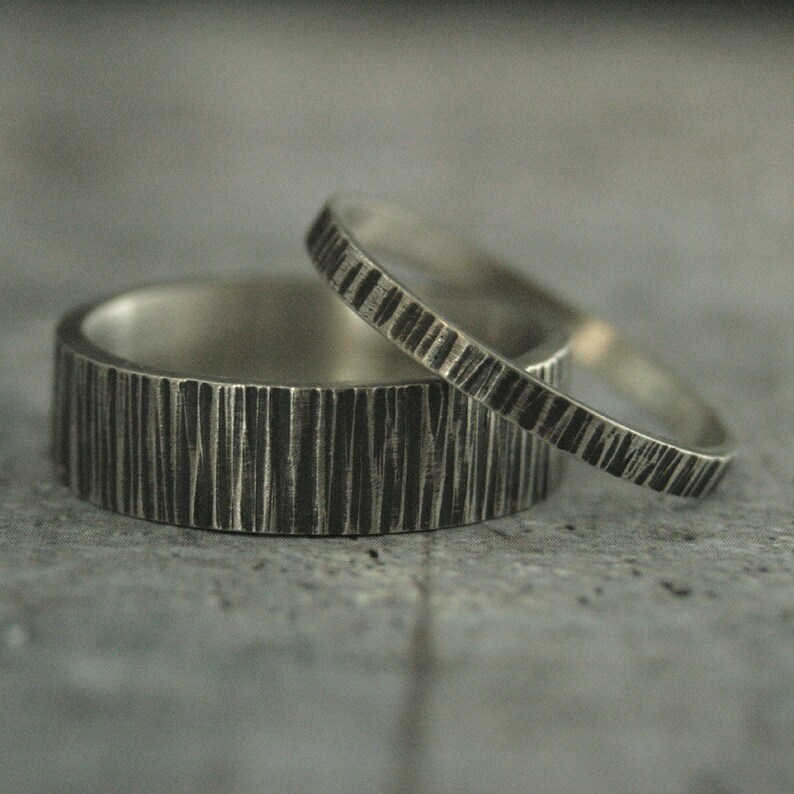 May include: Two silver rings with a textured, bark-like pattern. One ring is wider than the other, and both have a dark, oxidized finish. The rings are set against a gray background.