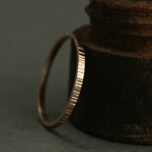 May include: A thin, rose gold band ring with a textured, hammered finish. The ring is shown on a dark brown wooden surface.
