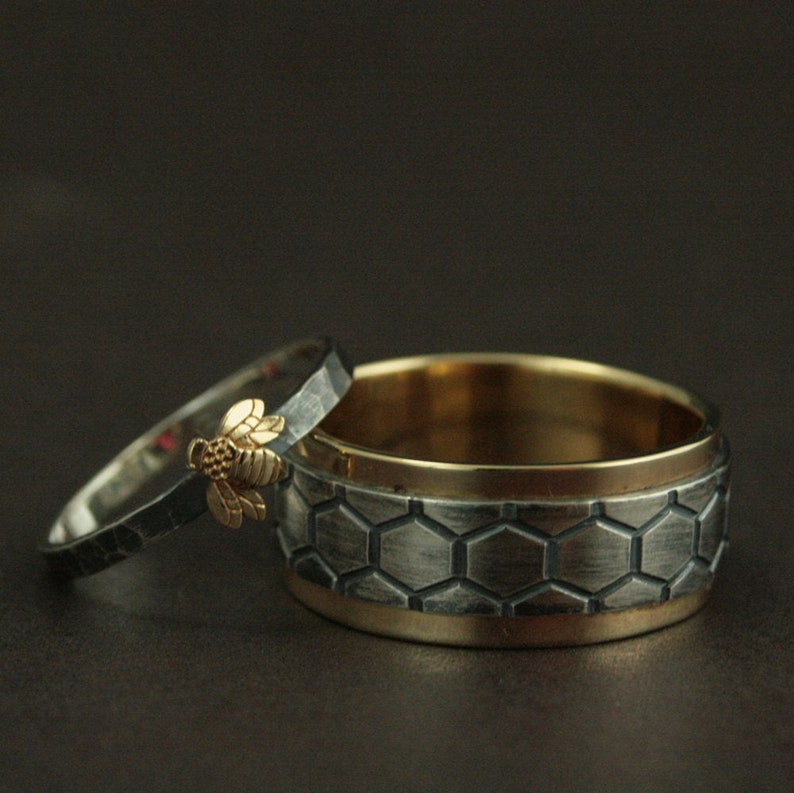 May include: Two wedding rings. One ring is silver with a gold bee accent. The other ring is silver with a honeycomb pattern and a gold interior. The rings are set against a dark background.
