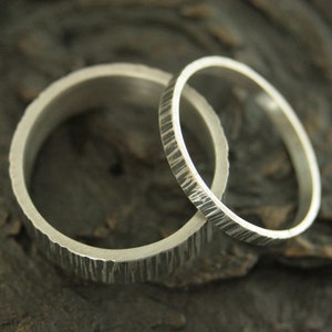 May include: Two silver wedding rings with textured surfaces. One ring is wider than the other. The rings are set against a dark, textured background, highlighting their metallic sheen and unique design.