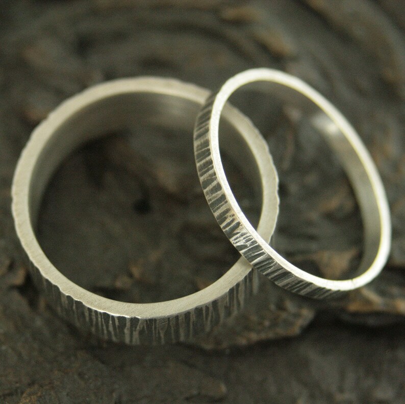 May include: Two silver wedding rings with textured surfaces. One ring is wider than the other. The rings are set against a dark, textured background, highlighting their metallic sheen and unique design.