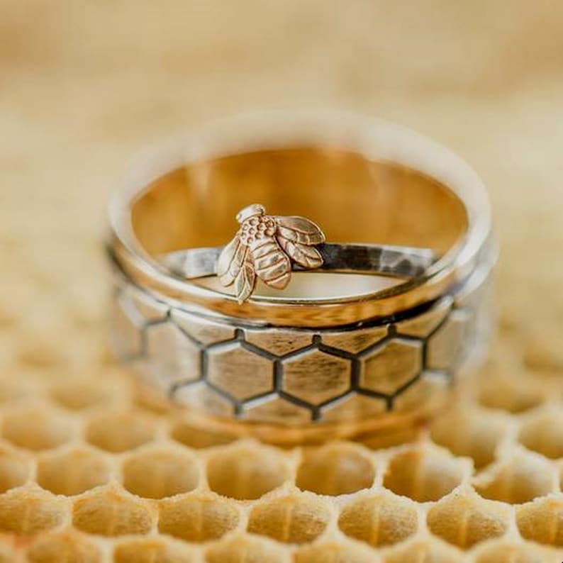 May include: A close-up of a gold and silver ring set. The top ring is gold, and the bottom ring is silver with a honeycomb pattern. A small gold bee is attached to the top ring. The rings are on a honeycomb surface.