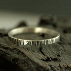 May include: A silver ring with a textured, bark-like pattern. The ring is thin and circular, with vertical lines etched around the band. The ring is displayed on a dark, textured surface, creating a contrast with the bright metal.