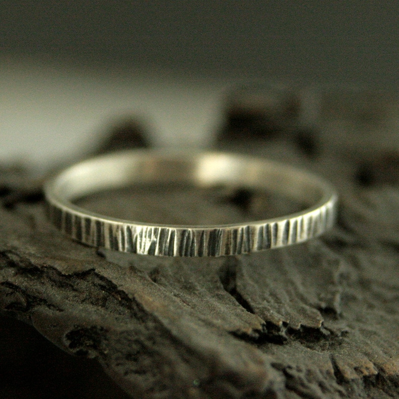 May include: A silver ring with a textured, bark-like pattern. The ring is thin and circular, with vertical lines etched around the band. The ring is displayed on a dark, textured surface, creating a contrast with the bright metal.