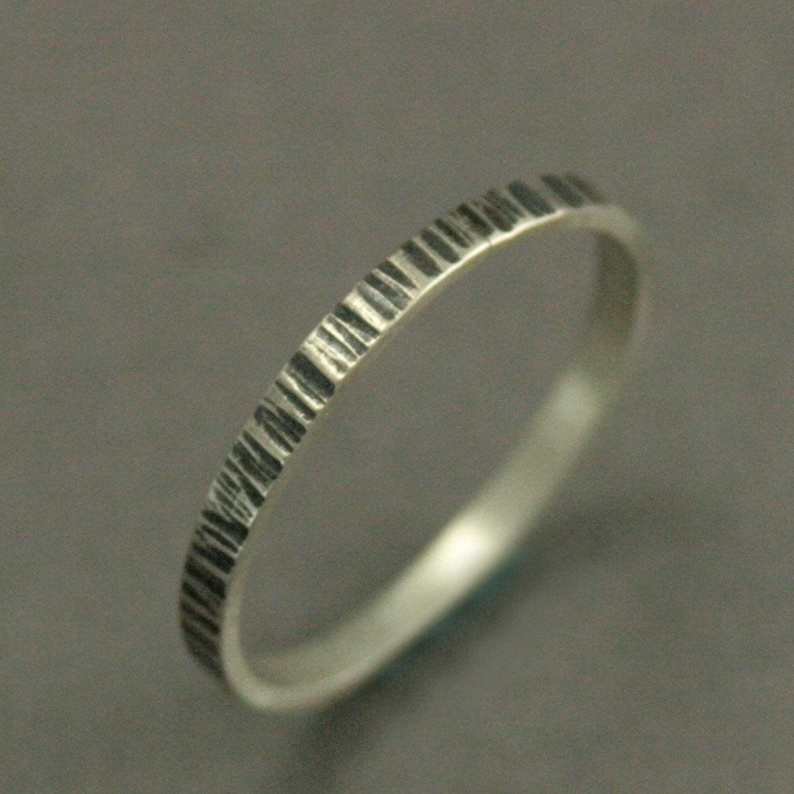 May include: A silver ring with a textured, bark-like pattern around the band. The ring has a simple, elegant design, perfect for everyday wear. The ring is shown against a gray background.