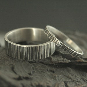 May include: Two silver rings with textured, vertical line patterns. One ring is wider than the other. The rings are displayed on a dark, textured surface, creating a contrast that highlights the metallic sheen.