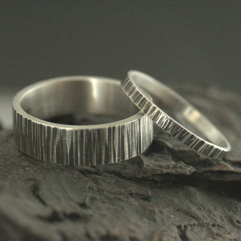 May include: Two silver rings with textured, vertical line patterns. One ring is wider than the other. The rings are displayed on a dark, textured surface, creating a contrast that highlights the metallic sheen.