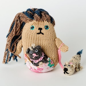 Peut inclure: Un jouet en tricot marron avec des cheveux en laine bleu et marron, tenant deux plus petits jouets en tricot marron avec des colliers roses. Le plus grand jouet porte une jupe en tissu rose et fleurie.