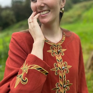 May include: Rust-colored jacket with ornate embroidery in gold, black, and brown. The jacket features detailed floral and geometric patterns around the neckline, cuffs, and down the front, with silver-colored buttons. The sleeves are long, and the fabric appears to be a textured material.
