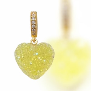 Puede incluir: Un dije dorado con un colgante en forma de corazón amarillo. El corazón está cubierto de cristales brillantes. La sección superior del dije es una barra dorada con pequeños cristales transparentes. El fondo está borroso, con una segunda forma de corazón visible.