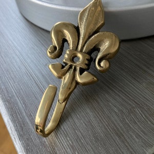 May include: A gold-toned metal hook with a fleur-de-lis design. The hook is attached to a wall or surface.