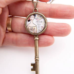 Può includere: Una collana con una chiave in bronzo e un ciondolo rotondo in vetro con un'illustrazione vintage di una donna con la parola "GOOD" e la frase "The things that make you happy" stampata su di esso.