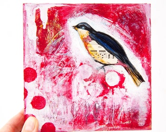 Puede incluir: Una obra de arte cuadrada de técnica mixta que presenta un pájaro con la espalda negra, el pecho amarillo y un ala hecha de partituras. El fondo es rojo texturizado con detalles en blanco y lunares rojos. La obra de arte está sostenida por una mano.
