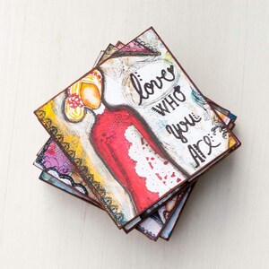 May include: A stack of colorful square coasters with a painted image of a woman in a red dress and the words "love who you are" on the top coaster.