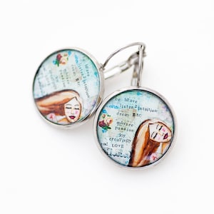 May include: A pair of silver hoop earrings with a round, blue and white glass charm. The charm features a watercolor illustration of a woman with long brown hair and the text "be brave listen 2 intuition dream BIG embrace passion joy creativity LOVE".