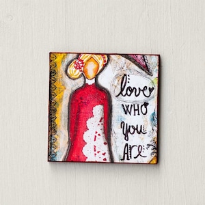 May include: A colorful painting on a wooden canvas featuring a woman with red hair and a red dress. The text "love who you are" is written in black paint.
