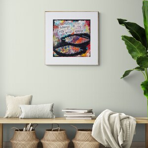 Puede incluir: Una impresi&oacute;n de arte enmarcada con un dise&ntilde;o abstracto colorido que presenta plumas y el texto "blame it on my wild spirit". La impresi&oacute;n est&aacute; colgada en una pared verde claro sobre un banco de madera con una manta blanca, dos cestas tejidas y dos almohadas blancas.