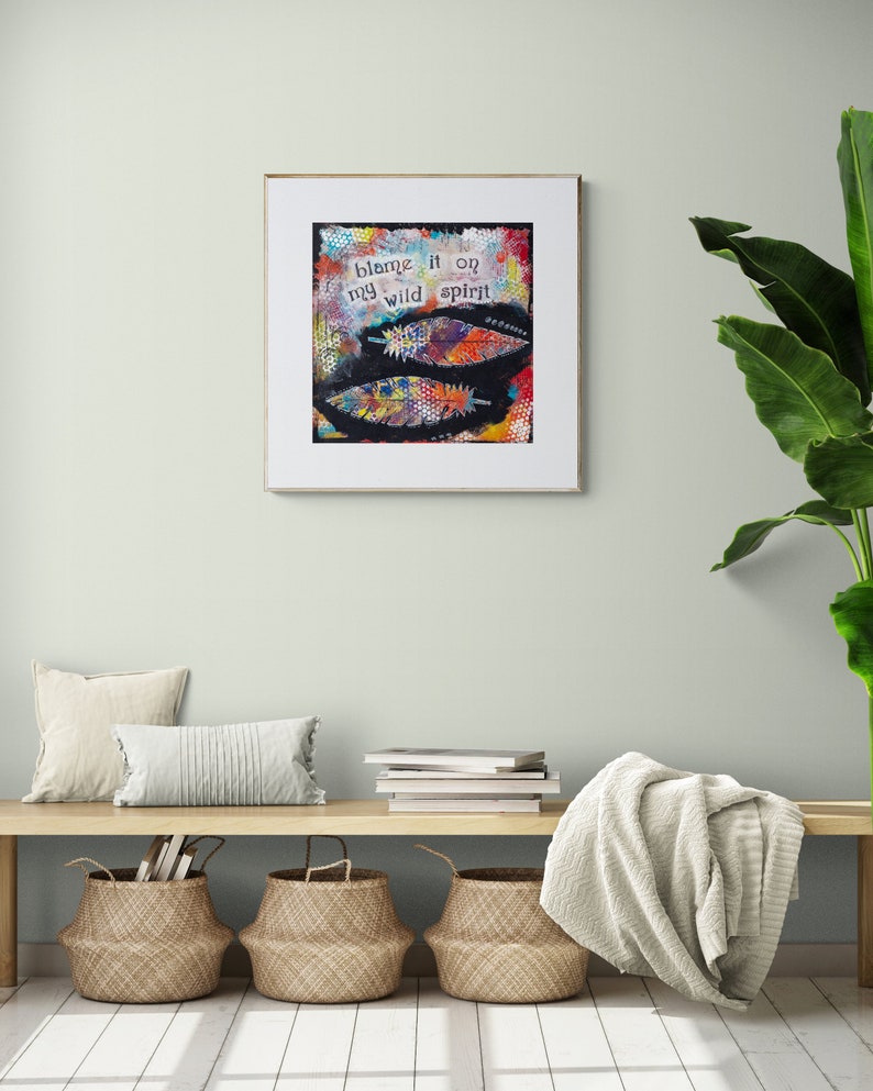Puede incluir: Una impresi&oacute;n de arte enmarcada con un dise&ntilde;o abstracto colorido que presenta plumas y el texto "blame it on my wild spirit". La impresi&oacute;n est&aacute; colgada en una pared verde claro sobre un banco de madera con una manta blanca, dos cestas tejidas y dos almohadas blancas.