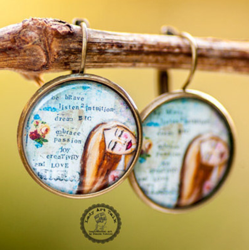 May include: A pair of round, bronze-colored earrings with a glass cabochon featuring a watercolor painting of a woman's face with flowers and the text "be brave, listen to intuition, dream big, embrace passion, joy, creativity and love".