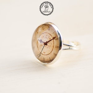 Pode incluir: Um anel de prata com um mostrador de rel&oacute;gio redondo de cor creme com algarismos romanos e as palavras "The Clock is Always Ticking".