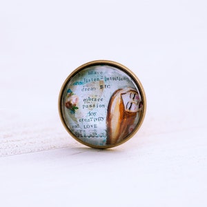 May include: A round glass cabochon with a gold metal frame. The cabochon features a watercolor illustration of a woman with long brown hair and a quote that reads: "be brave, listen to intuition, dream big, embrace passion, joy, creativity, and love."