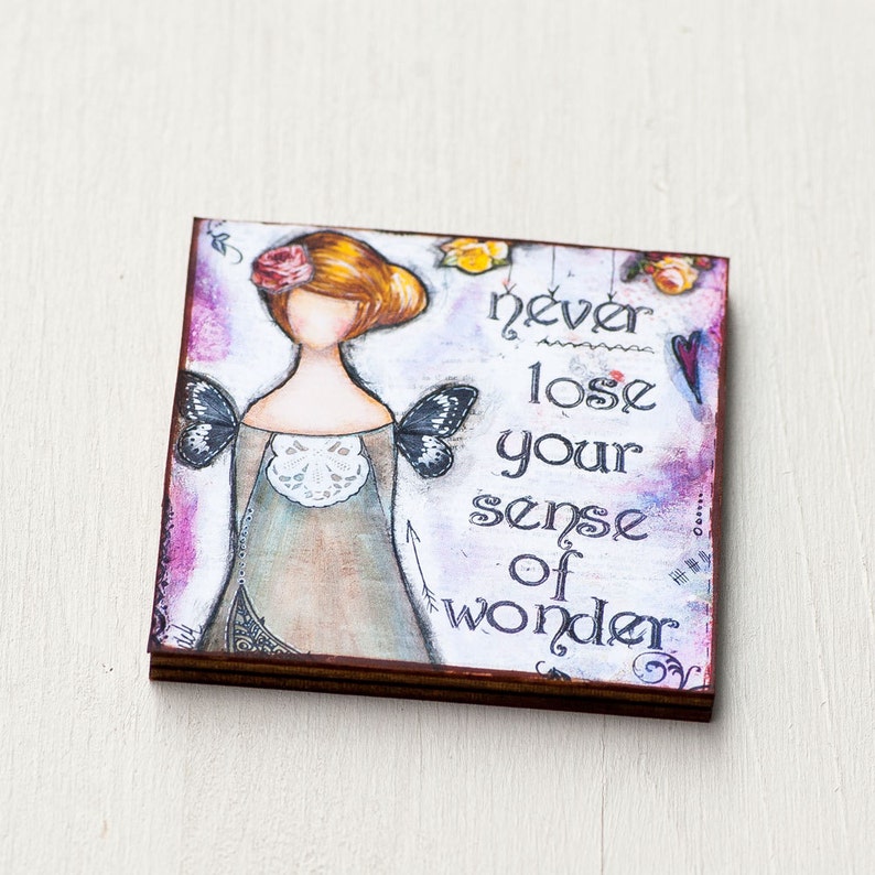 K&ouml;nnte beinhalten: Ein Aquarellbild auf einer Holztafel mit einer skurrilen Illustration eines M&auml;dchens mit Fl&uuml;geln und dem Text "never lose your sense of wonder".
