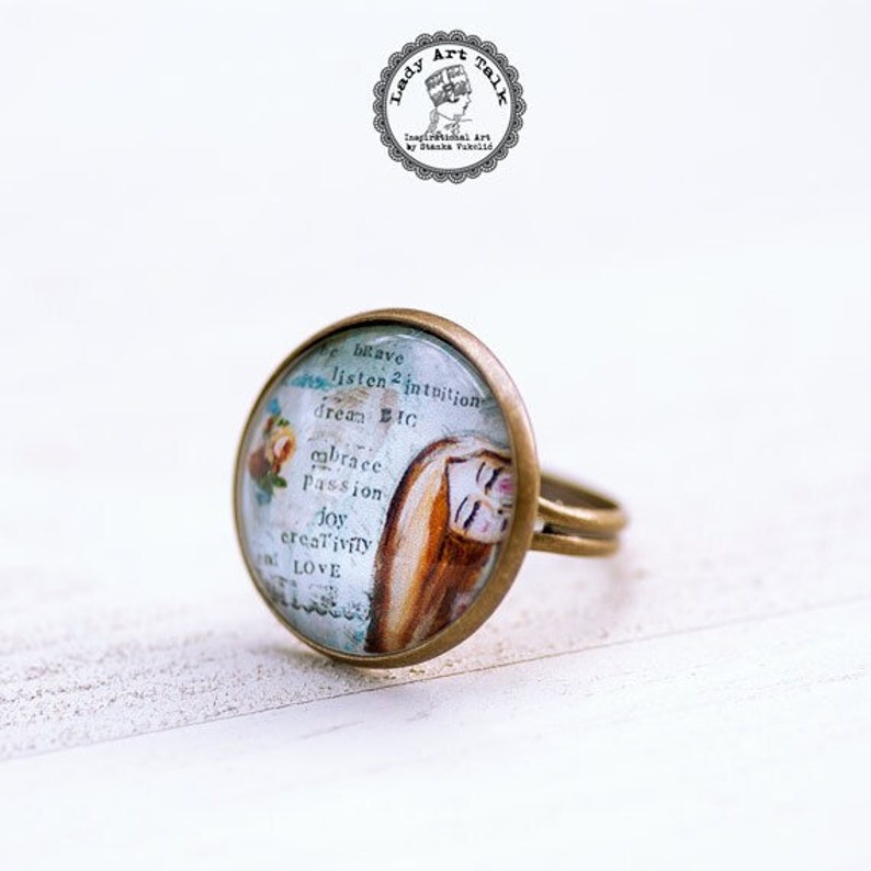 May include: A gold ring with a round glass cabochon featuring a watercolor painting of a woman's face and the words "believe, listen 2 intuition, dream etc, embrace passion, joy creativity, love".