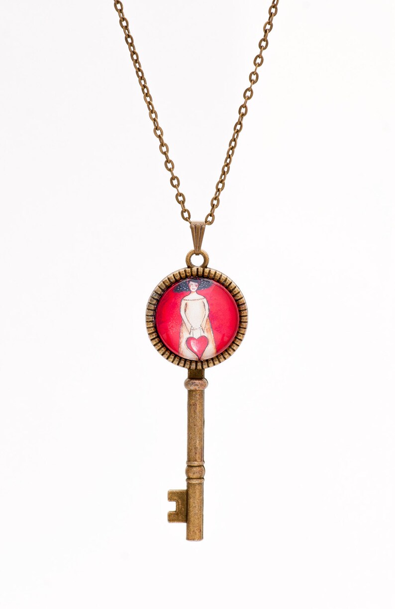 May include: A gold key necklace with a round pendant featuring a woman holding a red heart. The pendant is set against a red background.