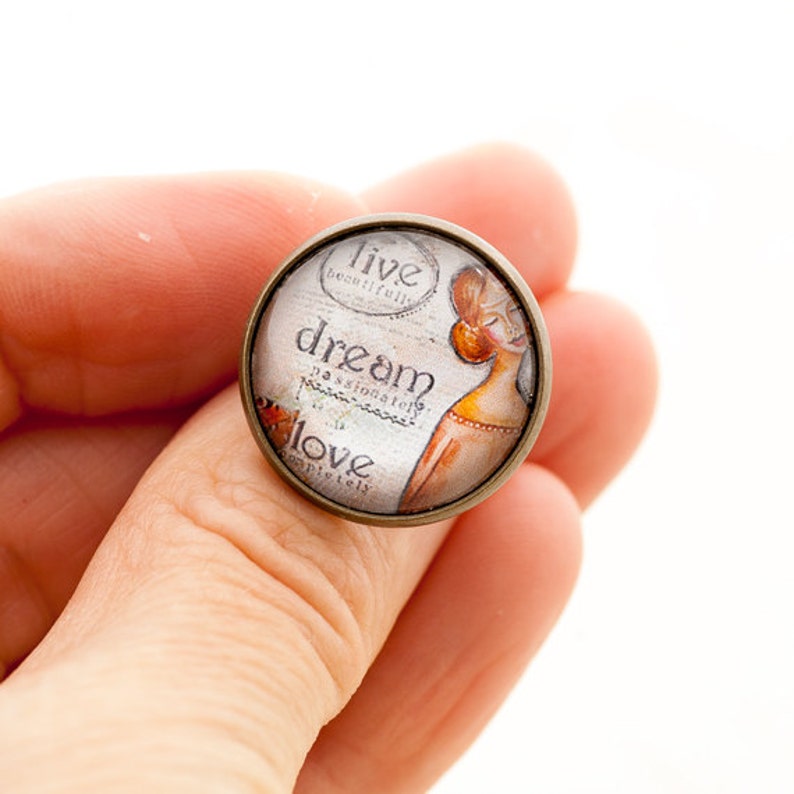Puede incluir: Un anillo redondo de vidrio con un engaste de bronce. El vidrio presenta una ilustraci&oacute;n de estilo vintage de una mujer con las palabras "live beautifully, dream passionately, love completely" impresas en el fondo.