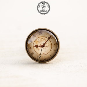 Pode incluir: Um bot&atilde;o redondo de cor lat&atilde;o com uma face de vidro que apresenta um design de rel&oacute;gio vintage. O mostrador do rel&oacute;gio tem algarismos romanos, um ponteiro dos minutos e um ponteiro das horas. O mostrador do rel&oacute;gio &eacute; de cor creme com acentos marrons e o texto "L'heure" em uma fonte cursiva.