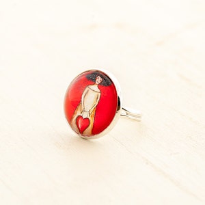 May include: A silver ring with a red and white cameo featuring a woman holding a red heart. The cameo is set in a round silver bezel.