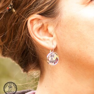 May include: Silver dangle earrings with a round charm that says "Life is Magic" in black script. The charm has a floral design with pink, yellow, and green accents.