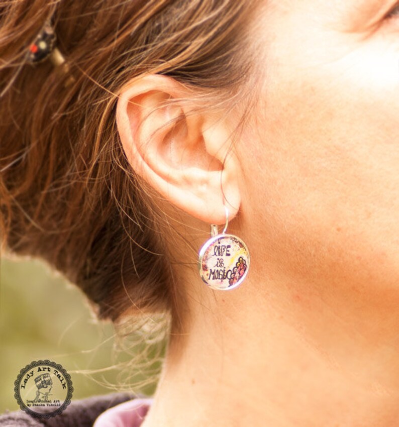 May include: Silver dangle earrings with a round charm that says "Life is Magic" in black script. The charm has a floral design with pink, yellow, and green accents.