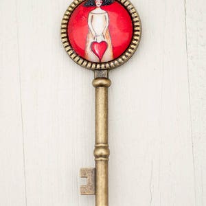 May include: A gold key necklace with a round pendant featuring a woman holding a red heart. The pendant has a red background and a gold frame.