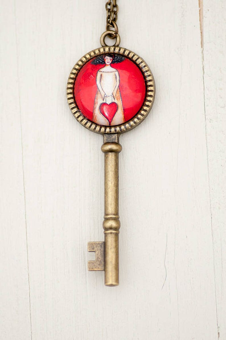 May include: A gold key necklace with a round pendant featuring a woman holding a red heart. The pendant has a red background and a gold frame.