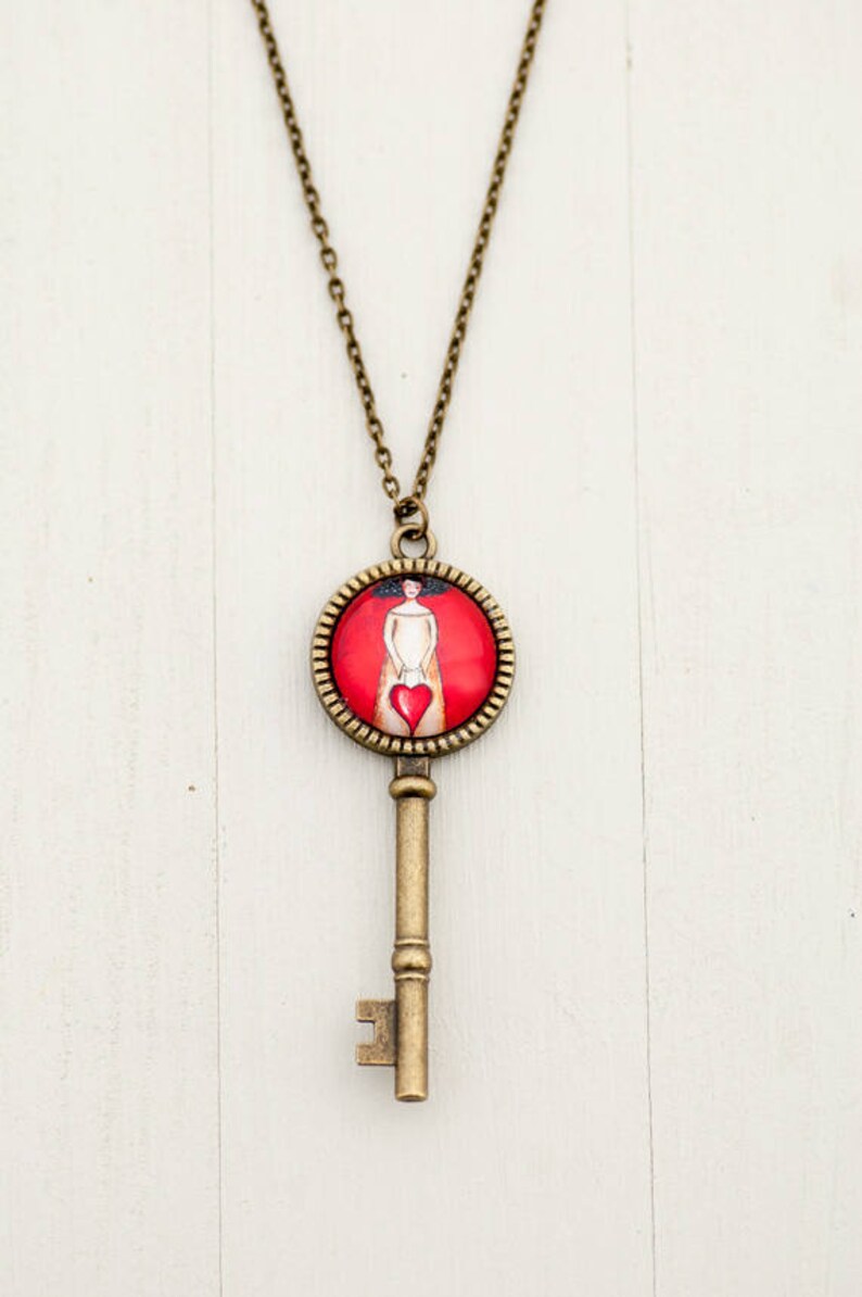 May include: A gold key necklace with a round pendant featuring a woman holding a red heart. The pendant is surrounded by a gold frame.