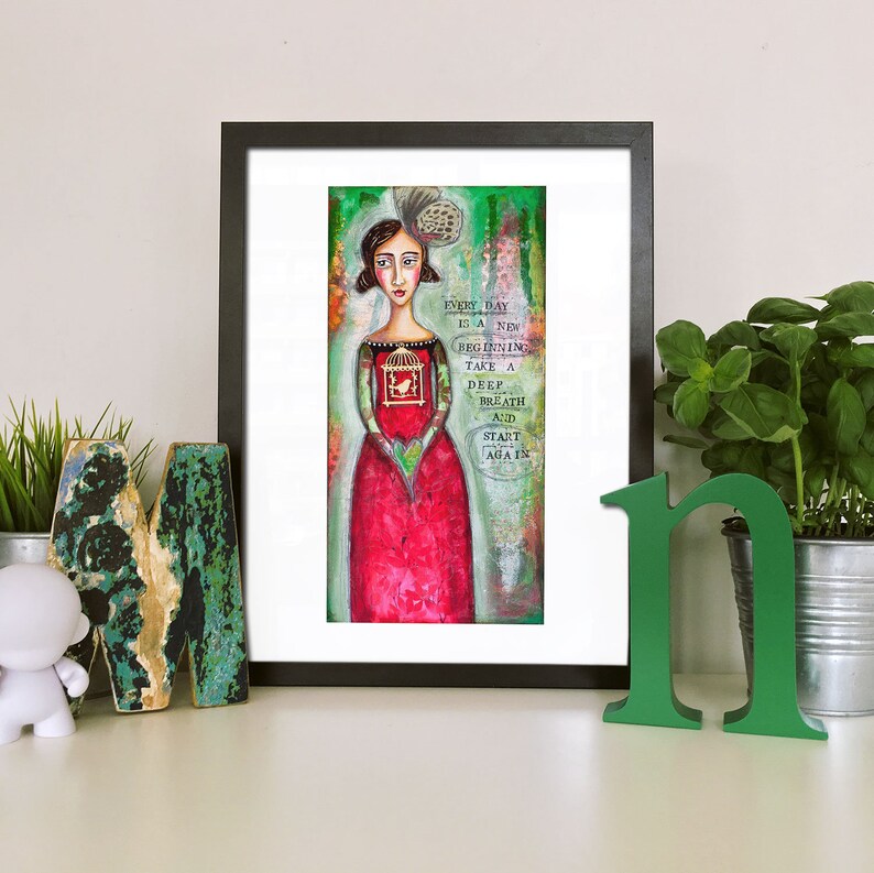 May include: A framed art print featuring a woman in a red dress holding a birdcage with a bird inside. The background is a green and brown abstract design. The text on the print reads "Every day is a new beginning. Take a deep breath and start again."