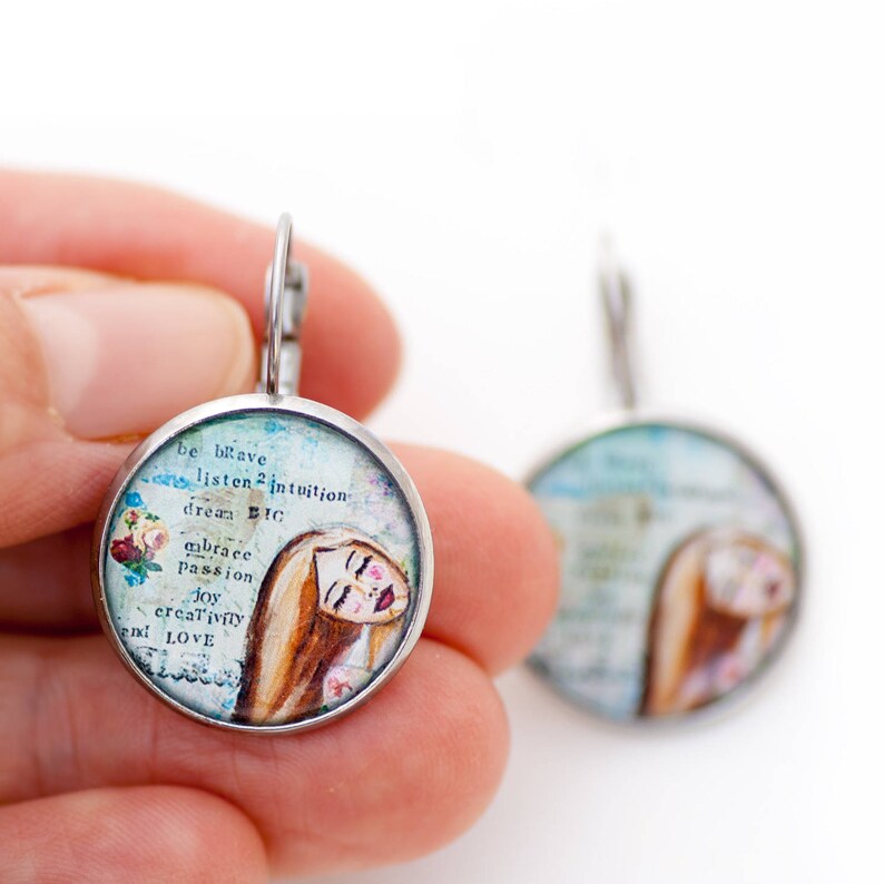 May include: A pair of silver earrings with a round, glass charm featuring a watercolor illustration of a woman with long brown hair and the text "be brave, listen to intuition, dream BIG, embrace passion, joy, creativity and LOVE".