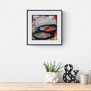 Puede incluir: Una impresi&oacute;n de arte enmarcada con un fondo abstracto colorido y el texto "blame it on my wild spirit". La impresi&oacute;n se muestra en una pared blanca con una planta en maceta y un signo de ampersand negro en frente.