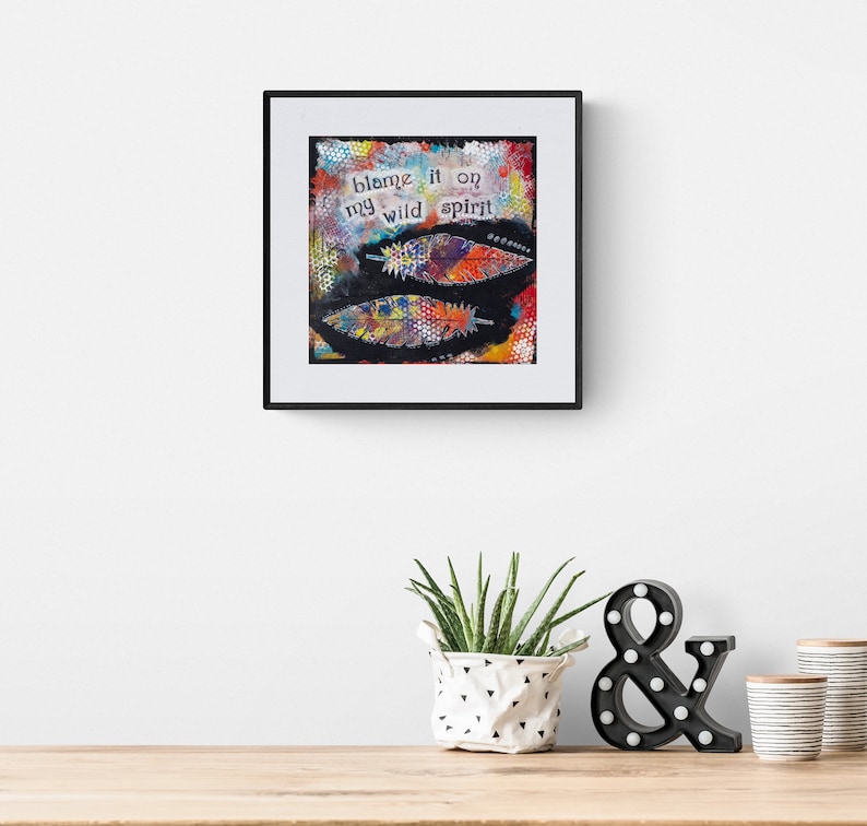Puede incluir: Una impresi&oacute;n de arte enmarcada con un fondo abstracto colorido y el texto "blame it on my wild spirit". La impresi&oacute;n se muestra en una pared blanca con una planta en maceta y un signo de ampersand negro en frente.