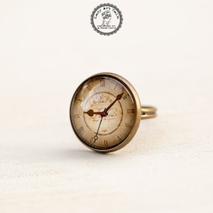 May include: A brass ring with a round, glass face featuring a vintage clock face with Roman numerals and the word "Histoire" in a decorative script.