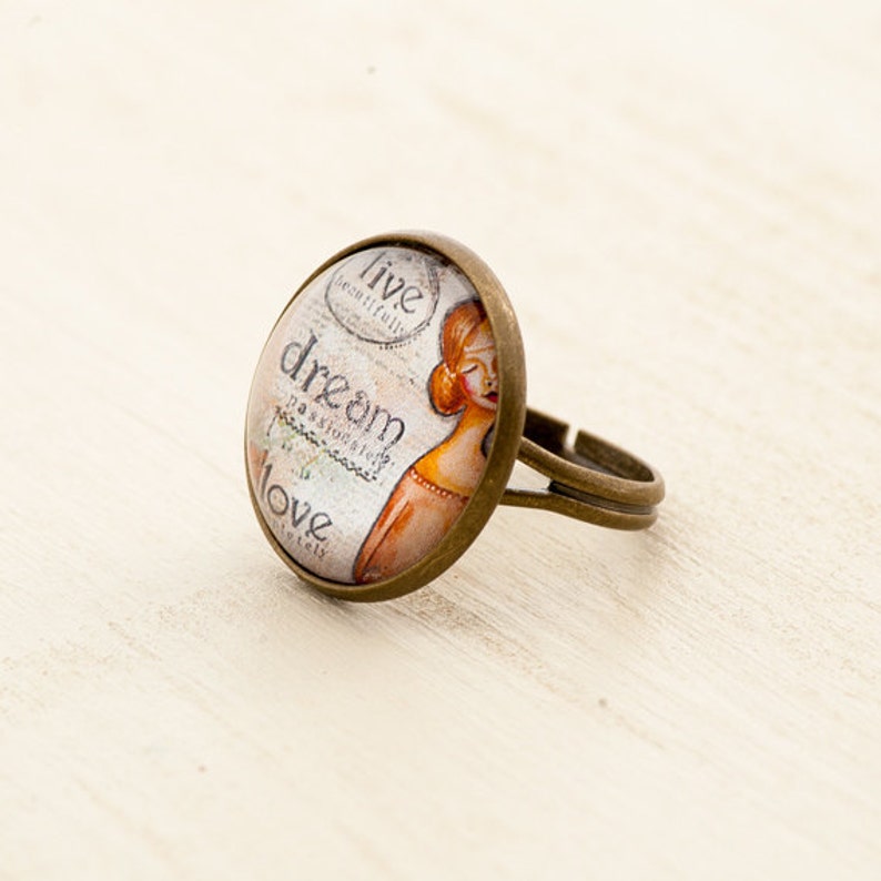 Puede incluir: Un anillo de bronce con un cabuj&oacute;n redondo de vidrio que presenta una ilustraci&oacute;n de estilo vintage de una mujer con las palabras "vive bellamente, sue&ntilde;a apasionadamente, ama completamente" impresas en &eacute;l.
