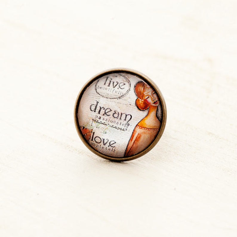 Puede incluir: Un cabuj&oacute;n redondo de vidrio con un engaste de bronce presenta una ilustraci&oacute;n de estilo vintage de una mujer con las palabras "live beautifully, dream passionately, love completely" impresas en el fondo.
