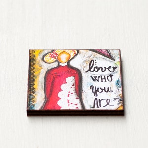 May include: A colorful painting on wood with a woman in a red dress and the words "love who you are" in black.