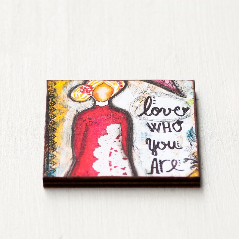 May include: A colorful painting on wood with a woman in a red dress and the words "love who you are" in black.