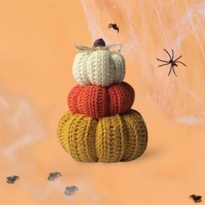 May include: A stacked decorative object consisting of three crocheted pumpkins in white, orange, and mustard yellow. The top white pumpkin features a small brown stem and a thin twine bow. Several small black plastic spiders are scattered around.