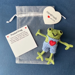 May include: A green crocheted frog wearing blue dungarees with a red heart on the front. The frog is holding its arms out and has a happy expression on its face. The frog is packaged in a white organza bag with a white heart-shaped tag that says "Handmade with love".